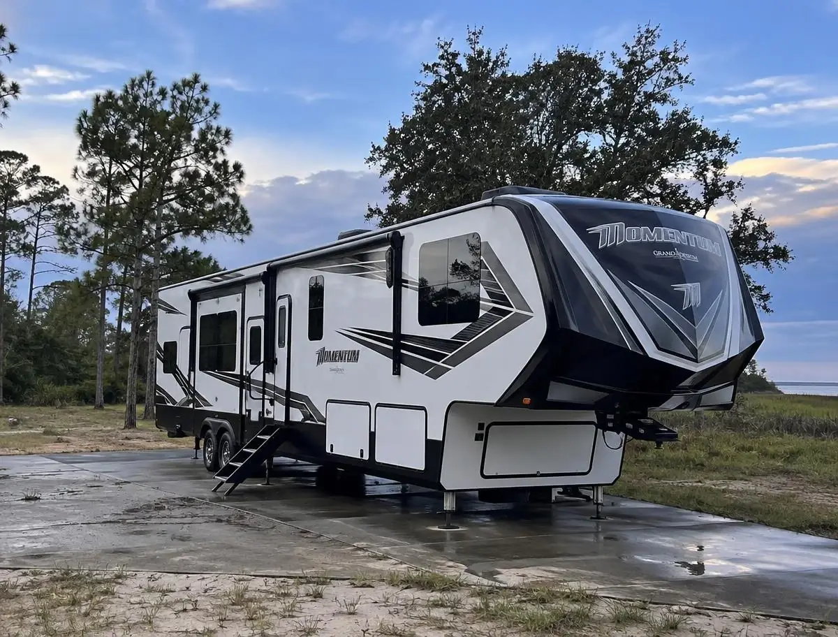 Fifth-wheel toy hauler parked on a wet residential driveway with puddles beneath a partly cloudy sky and late-day sunlight