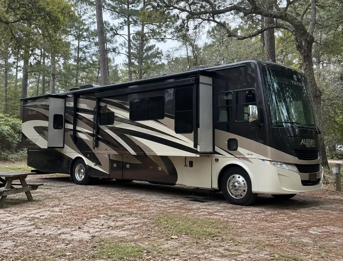 Class A motorhome with a slide-out extended and awning open at a sandy campsite under loblolly pines on a sunny day